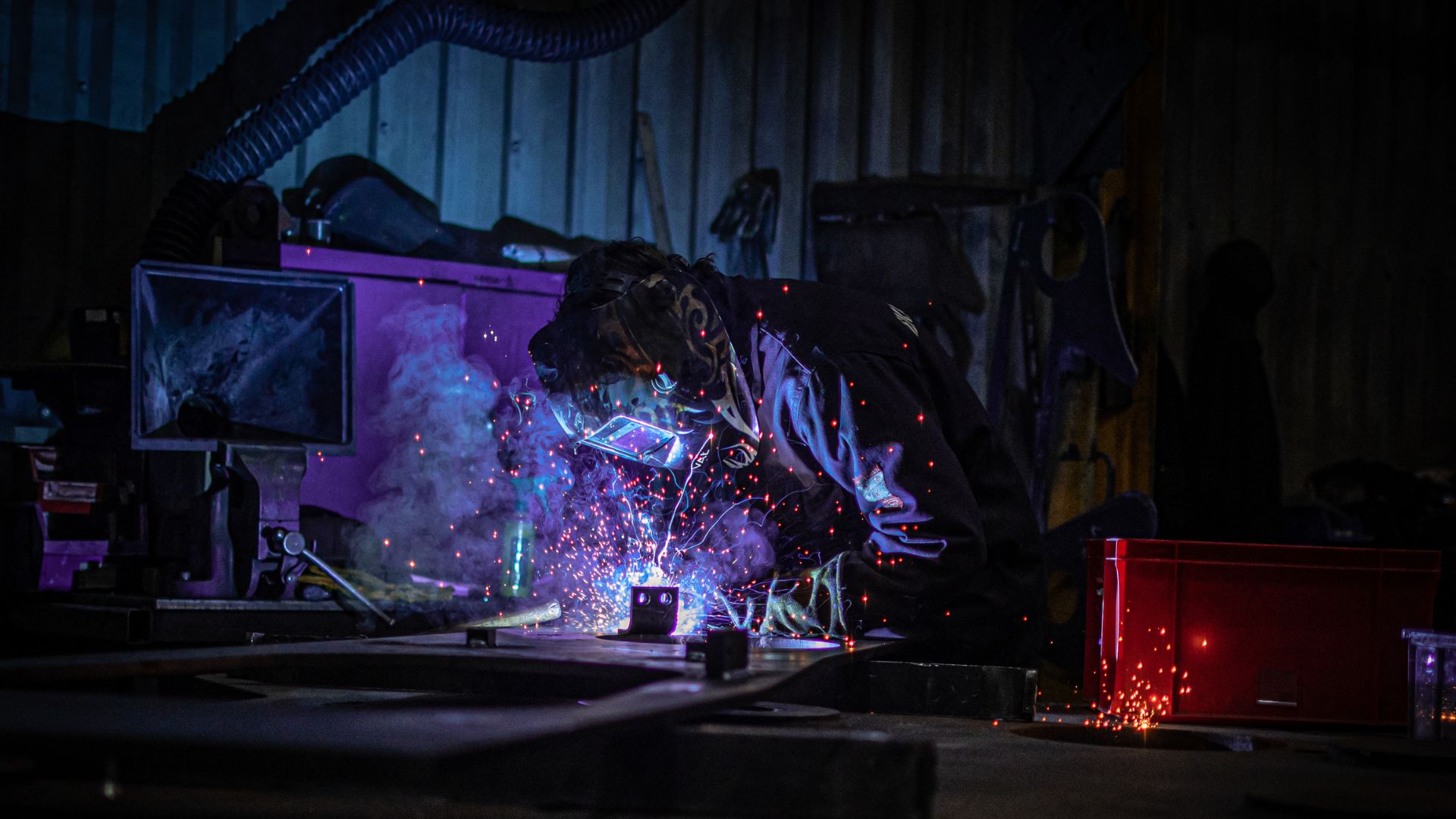 Person wearing welding mask and boiler suit welding a piece of metal with sparks flying.