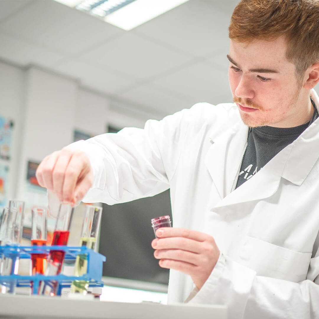 Young man in a white lab coat sat at a desk that has many beakers with coloured liquids on it.