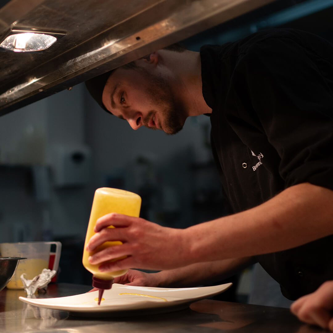 Chef holding a pot and spoon and dishing out food onto a plate.